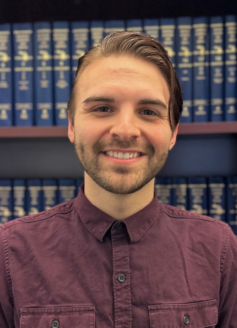 A young man with short brown hair, a trimmed beard, and a maroon button-up shirt smiles in front of a bookshelf filled with large blue books.